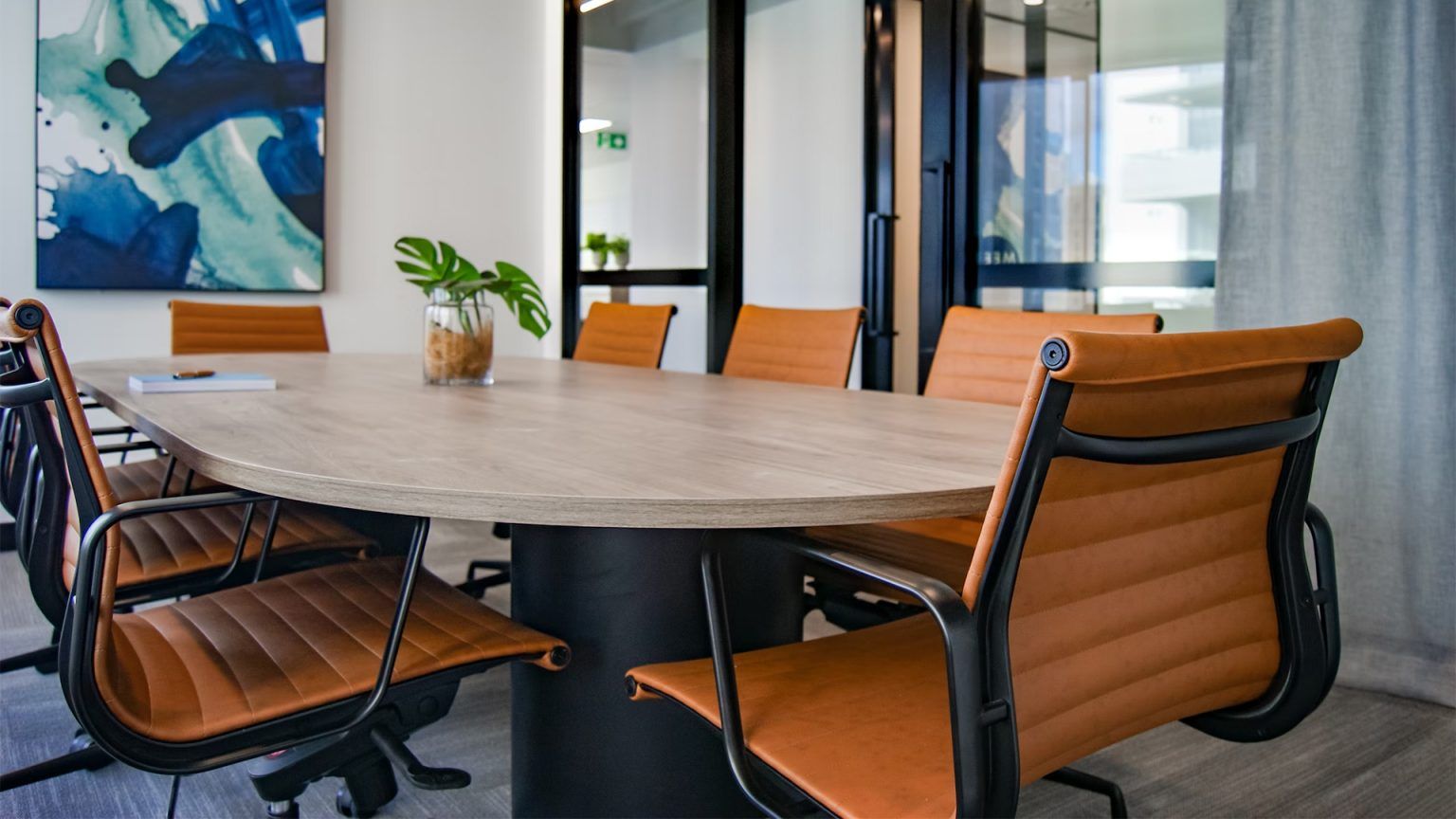  A conference room with a large wooden table and brown leather chairs, with a painting on the wall and a plant on the table, suitable for use as an immersive Zoom background.